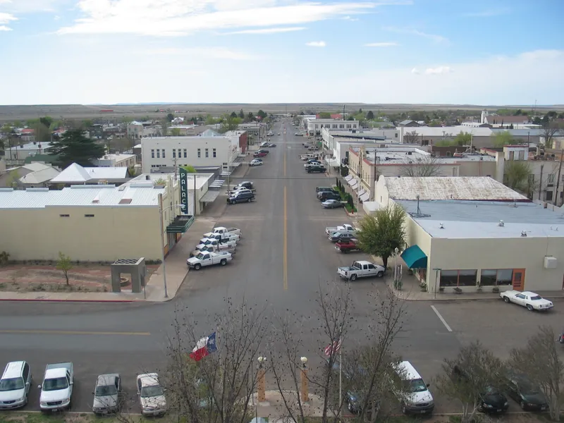 Looking down a wide main street of low buildings and parked pickups from a high window, with flat country beyond the town