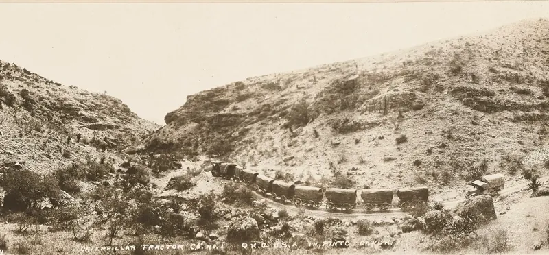 A long train of tracked tractors and wagons winding along a canyon floor between steep rocky slopes