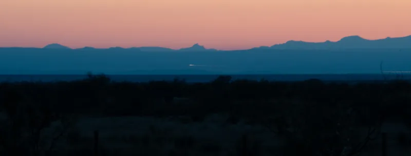Dusk from the Marfa Lights Viewing Area: the Chinati Mountains in silhouette against a pink sky, a dark blue band of distant flat below them with two short streaks of light, and dark shrubs in the foreground